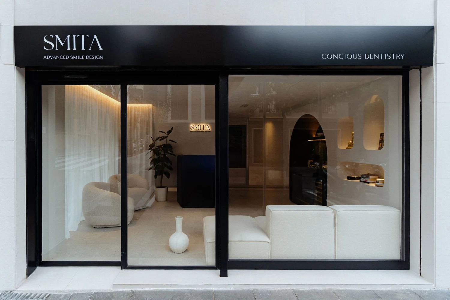 Smitasmile modern dental clinic storefront in Barcelona with large glass windows, black signage reading 'SMITA - Advanced Smile Design' and 'Conscious Dentistry', professional facade