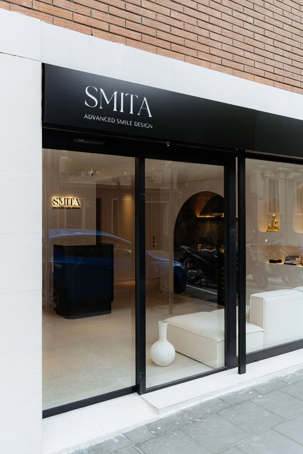 Smitasmile Barcelona entrance showing black signage, glass doors with beige contemporary seating, white interior accents, modern minimalist design reflecting Advanced Smile Design philosophy